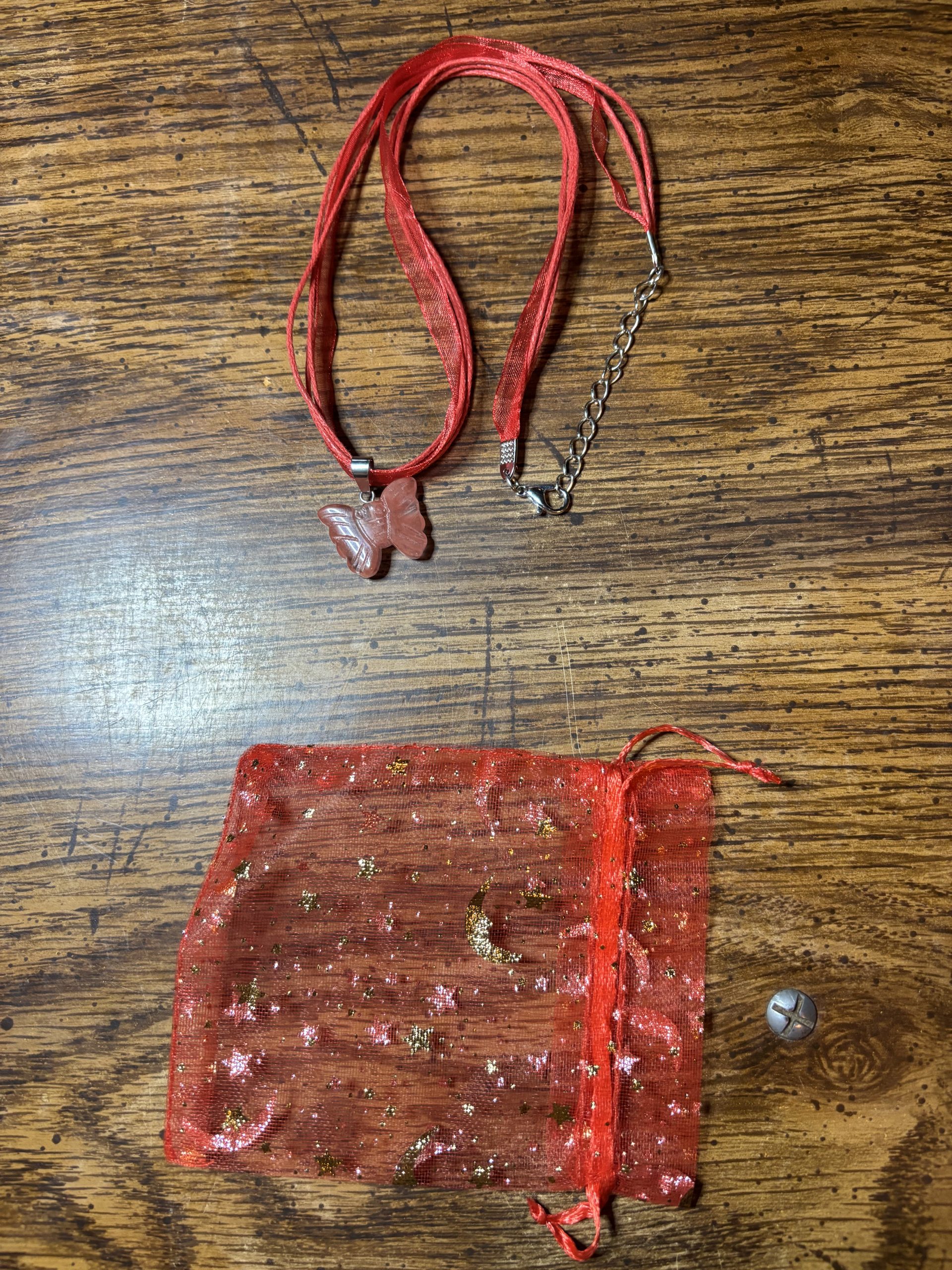 Red ribbon necklace with a butterfly pendant resting on a wooden table, beside a sparkly red gift pouch and metal chain.