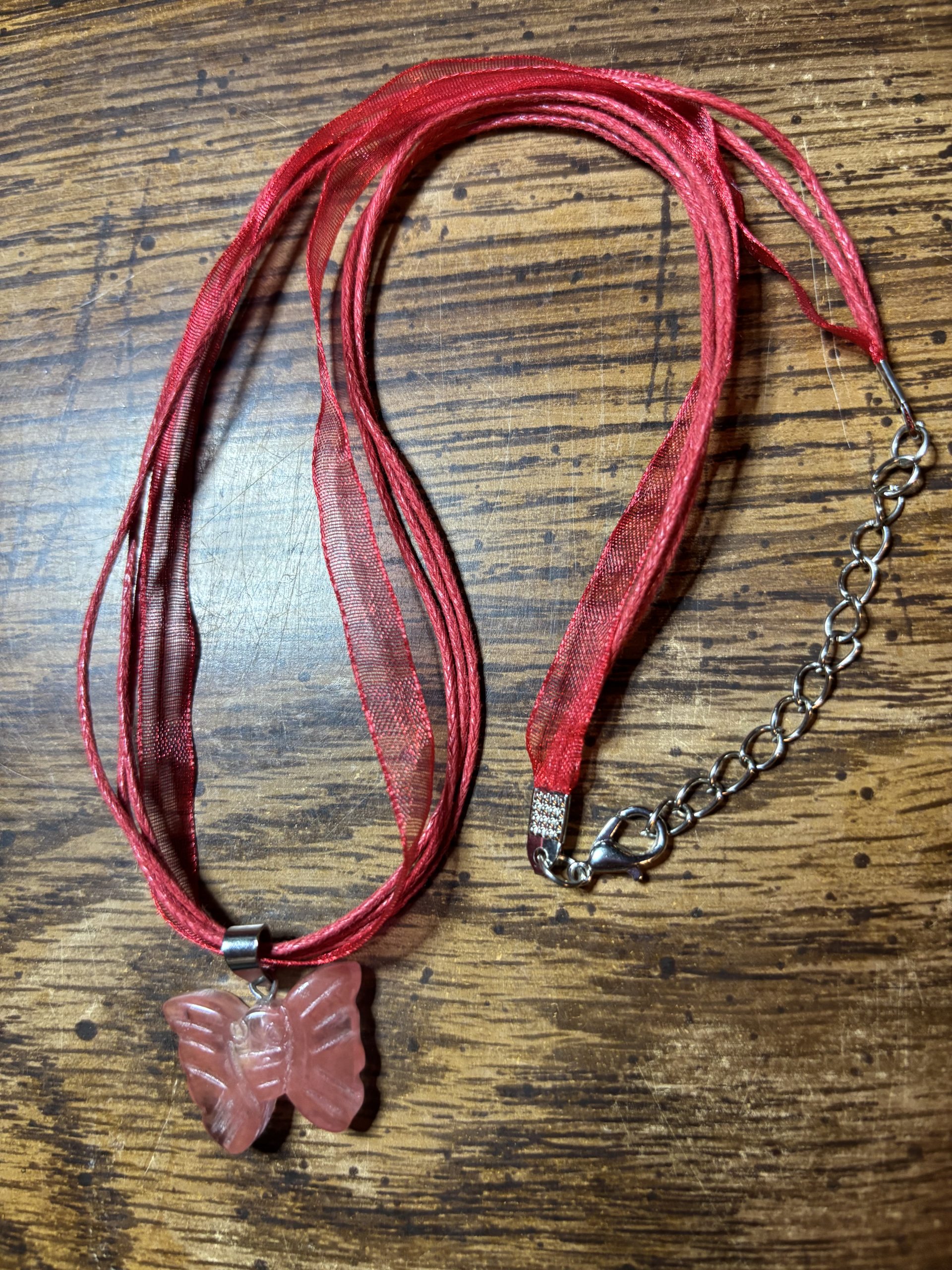 Red multi-strand ribbon necklace with a pink butterfly pendant on a metal chain resting on a wooden surface.